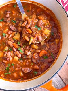 a large pot filled with beans and meats on top of a wooden table next to a spoon