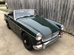 an old green car is parked in front of a wooden fence with a black top