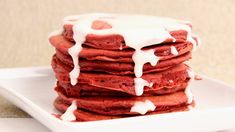 a stack of red velvet pancakes covered in white icing on a square white plate