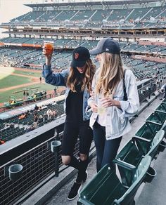 two women standing in the bleachers at a baseball game, one holding up a drink