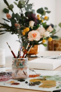 a jar filled with paint and brushes sitting on top of a table next to flowers