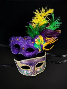 two masquerade masks with feathers and ribbons on a black background, one is purple the other is yellow