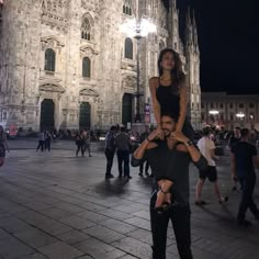 a man carrying a woman on his shoulders in front of a cathedral at night with people walking around