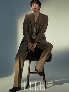 a man sitting on top of a wooden chair next to a white wall and wearing a suit