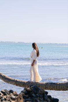 a woman in white dress walking across rocks near the ocean with her surfboard behind her