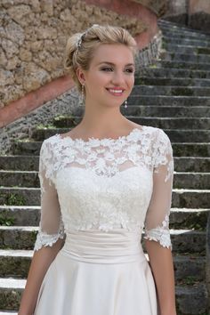 a woman wearing a wedding dress with sheer sleeves and lace on the top, standing in front of some stairs