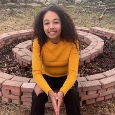 a young woman sitting in front of a brick fire pit with her hands folded out