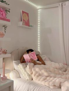 a woman laying in bed reading a book with lights on the wall above her head