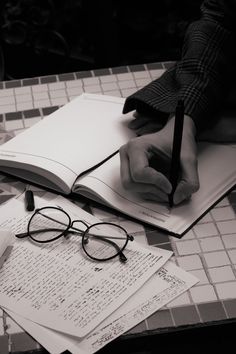 a person wearing glasses writing on a book with a pen and eyeglasses next to it