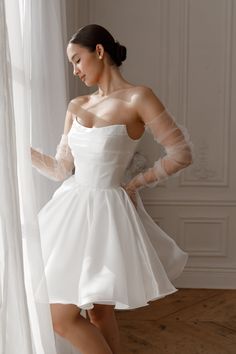 a woman in a short white dress standing next to a window