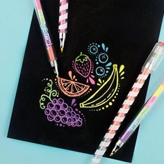 two pens are sitting on top of a black paper with fruit and berries drawn on it