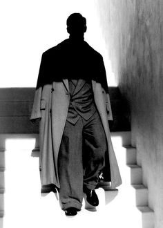 a man is walking down the stairs wearing a long coat and pants with his hands in his pockets