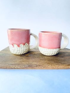 two coffee mugs sitting on top of a wooden board with pink and white glaze