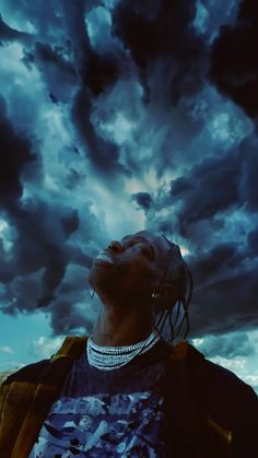 a man with dreadlocks standing in front of a cloudy sky and looking up at the sky