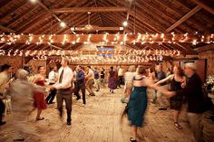 a group of people dancing in a barn with lights strung from the ceiling and wooden floors