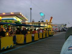 people are riding on a toy train at an amusement park in front of the boardwalk