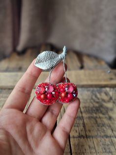 a hand holding two red heart shaped earrings on top of a wooden table next to a leaf