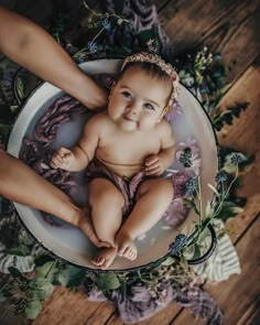 a baby is sitting in a bowl surrounded by flowers and greenery with her mother's hands