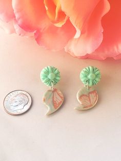 two green and pink flower shaped earrings next to a penny on a white surface with a rose in the background