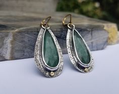 "I was drawn to aventurine because of its wonderful rich green coloring. I love the mossy green inclusions and the subtle sparkle of mica. These tiny flecks of mica add depth and interest to the stone. These earrings are sawn out of sterling silver sheet that has been textured with a pebble pattern. The long-faceted drops of aventurine have been bezel set in fine silver. The backs of the earrings have been sawn out to allow light to enter from the back illuminating the stone. A disk of 18K gold Elegant Green Earrings With Natural Stones, Elegant Green Hammered Earrings, Green Moss Agate Earrings For Gift, Green Artisan Gemstone Earrings, Artisan Green Gemstone Earrings, Elegant Green Patina Earrings, Green Multi-stone Sterling Silver Earrings, Gold Color Combination, Designer Board