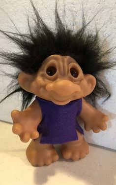 a troll doll with long black hair on it's head and purple shirt in front of a white wall