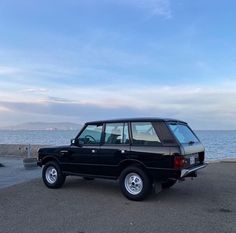 an old black car parked on the side of the road next to the ocean with mountains in the background