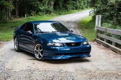 a blue sports car parked on the side of a dirt road next to a forest