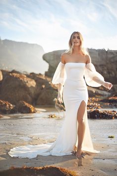 a woman standing on top of a sandy beach wearing a white dress and holding her arms out