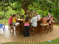 a group of people sitting around a wooden table in the middle of a park area