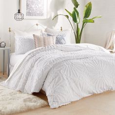 a bed with white comforter and pillows in a room next to a plant on the floor