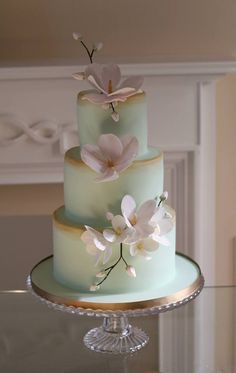 a three tiered cake with white flowers on the top and green frosting, sitting on a glass table