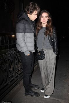 a man and woman standing next to each other on a sidewalk at night with the lights on