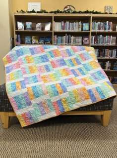 a multicolored quilt sitting on top of a bench in front of a bookcase