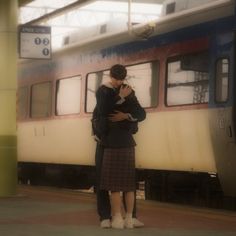 two people standing next to each other in front of a train at a station with their arms around each other