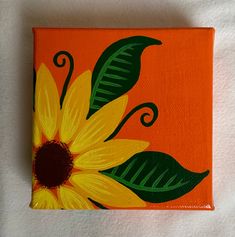 an orange and yellow painted square with green leaves on the bottom is sitting on a white surface