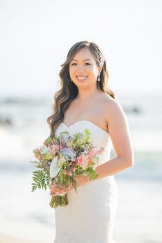 a woman in a wedding dress is holding a bouquet on the beach and smiling at the camera