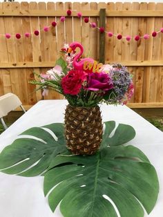 a pineapple vase filled with flowers and greenery on a white table cloth in front of a wooden fence