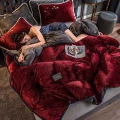 a woman laying on top of a red bed covered in pillows and blankets next to a lamp