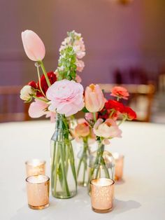 vases filled with pink and red flowers sit on a white tablecloth, surrounded by candles