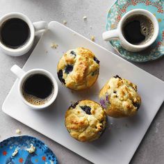 three blueberry muffins on a plate with two cups of coffee next to them