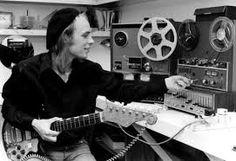 black and white photograph of man playing guitar in front of sound board with tape recorder