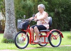 an older woman riding on the back of a red bike in a park with trees