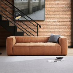 a brown couch sitting in front of a brick wall next to a stair case and lamp