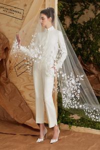 a woman standing in front of a wooden structure wearing a white suit and veil with flowers on it