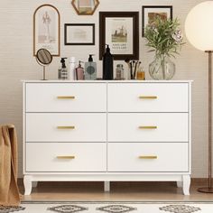 a white dresser topped with lots of drawers next to a lamp and pictures on the wall