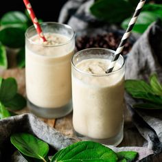 two glasses filled with smoothie on top of a wooden table next to green leaves