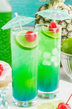 two glasses filled with green and blue liquid next to pineapples, cherries, and an umbrella