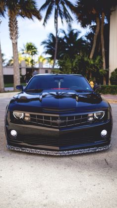 the front end of a black car parked in a parking lot with palm trees behind it