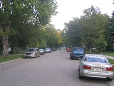 cars are parked on the street in front of some trees and grass, while another car is driving down the road