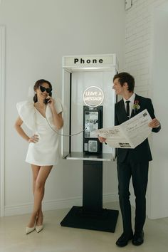 a man and woman standing in front of a phone booth holding papers while talking on the phone